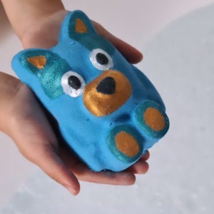 May include: A blue bath bomb shaped like a dog with gold and green accents. The dog has a black nose and white eyes.