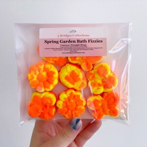 May include: Six orange and yellow bath fizzies in the shape of flowers. The bath fizzies are packaged in a clear plastic bag with a label that reads "Spring Garden Bath Fizzies" and "Fragrance: Pineapple Mango".