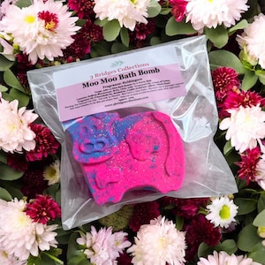 May include: A pink and blue cow-shaped bath bomb in a clear plastic bag, labelled "Moo Moo Bath Bomb." The bath bomb is surrounded by white and burgundy flowers. The label includes the fragrance "Bubblegum."