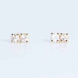 May include: A pair of gold stud earrings, each featuring two small, round, clear faceted stones set side-by-side in a prong setting. The metal has a polished yellow gold finish, and the stones are arranged horizontally on each earring post.