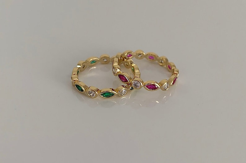 May include: Two gold rings with a row of small, clear, round stones and alternating green and pink marquise-shaped stones.