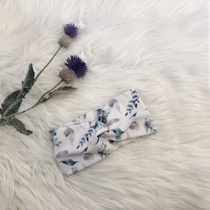 Könnte beinhalten: Ein weißes Stirnband mit einem Blumenmuster aus aquarellierten Blättern und Blüten in Violett- und Grüntönen. Das Stirnband ist verdreht und zu einem Knoten gebunden.