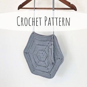 Könnte beinhalten: Eine graue gehäkelte Tasche in Hexagonform mit einem Quastenanhänger am Boden. Die Tasche hängt an einem Holzbügel mit dem Text "CROCHET PATTERN" darüber.