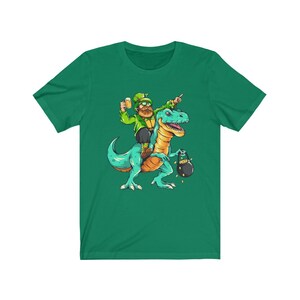 Puede incluir: Camiseta verde con una ilustración de dibujos animados de un duende montando un dinosaurio verde. El duende lleva una cerveza y una olla de oro.