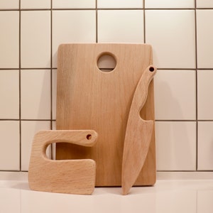 May include: A set of three wooden kitchen tools, including a cutting board, a knife, and a small utensil with a handle. The tools are made of light brown wood and have a natural finish.