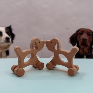 Può includere: Due cani giocattolo di legno che si guardano. I cani sono di colore marrone chiaro e hanno delle ruote ai piedi. I cani sono su una superficie blu chiaro.