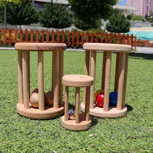 K&ouml;nnte beinhalten: Drei h&ouml;lzerne Rollspielzeuge mit unterschiedlich gro&szlig;en Zylindern. Der gr&ouml;&szlig;te Zylinder hat zwei Holzkugeln im Inneren, der mittlere Zylinder hat eine Glocke im Inneren und der kleinste Zylinder hat zwei farbige Kugeln im Inneren.