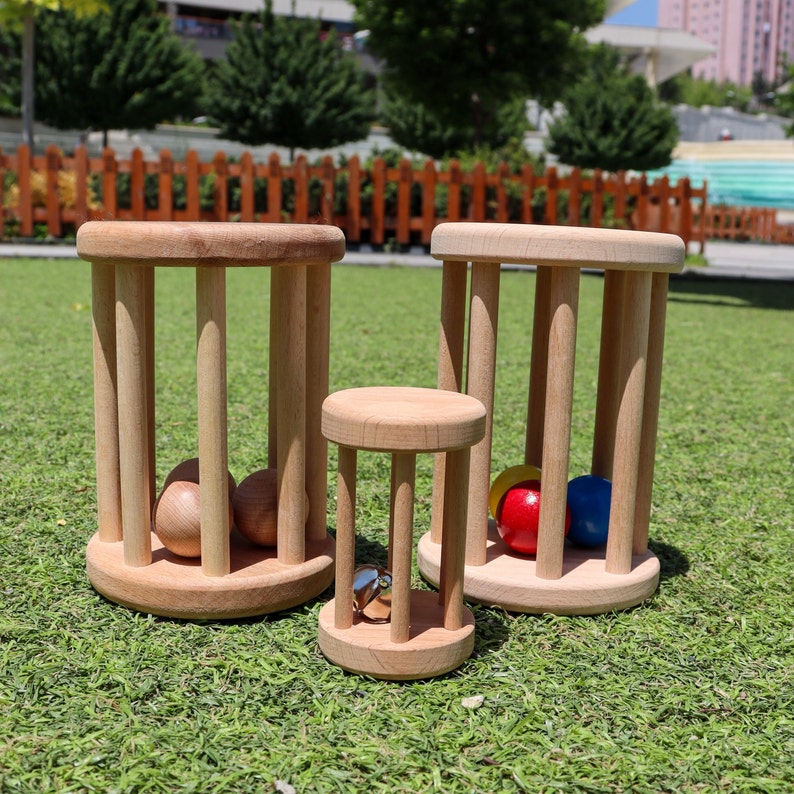 K&ouml;nnte beinhalten: Drei h&ouml;lzerne Rollspielzeuge mit unterschiedlich gro&szlig;en Zylindern. Der gr&ouml;&szlig;te Zylinder hat zwei Holzkugeln im Inneren, der mittlere Zylinder hat eine Glocke im Inneren und der kleinste Zylinder hat zwei farbige Kugeln im Inneren.