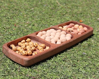 May include: A wooden serving tray with three compartments, filled with different types of nuts. The tray is made of light brown wood and has a natural finish. The nuts are arranged in a neat and organised manner.
