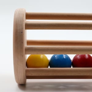 K&ouml;nnte beinhalten: Holzspielzeug mit zylindrischer Form und einer Reihe von L&ouml;chern. Drei farbige Kugeln, gelb, blau und rot, befinden sich im Inneren des Zylinders.