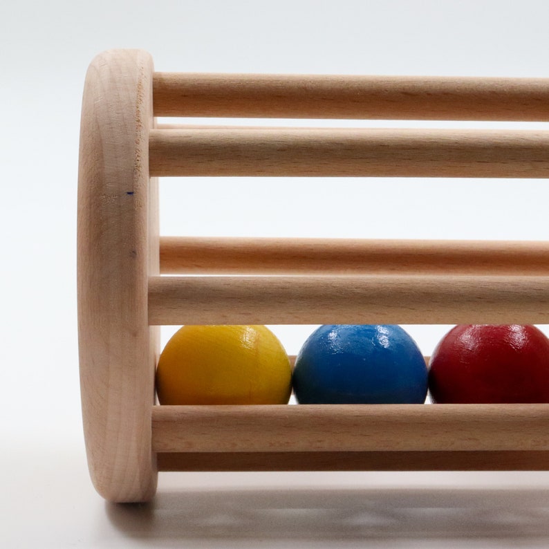 K&ouml;nnte beinhalten: Holzspielzeug mit zylindrischer Form und einer Reihe von L&ouml;chern. Drei farbige Kugeln, gelb, blau und rot, befinden sich im Inneren des Zylinders.