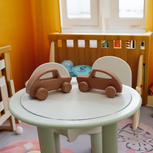 Könnte beinhalten: Zwei hölzerne Spielzeugautos in hellbrauner Farbe stehen auf einem weißen Tisch mit einem hellgrünen Rand. Der Tisch steht in einem Raum mit einem Holzbett im Hintergrund.
