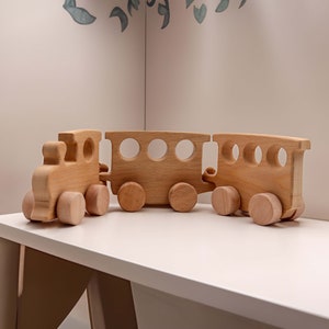 May include: A wooden toy train set with a locomotive and two carriages. The train is made of natural wood and has a light brown colour. The train is on a white surface.