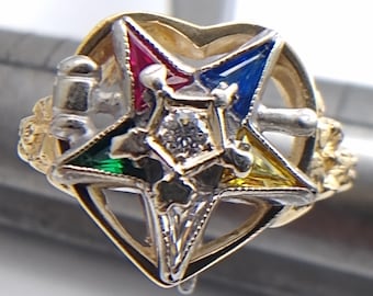 May include: A gold-toned ring featuring a multi-colored star design with a central diamond. The star is set within a heart-shaped frame. The ring has intricate detailing and a polished finish.
