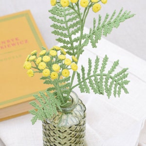 May include: A crocheted bouquet of yellow flowers with green leaves in a clear, textured glass vase. The vase is sitting on a white linen surface.