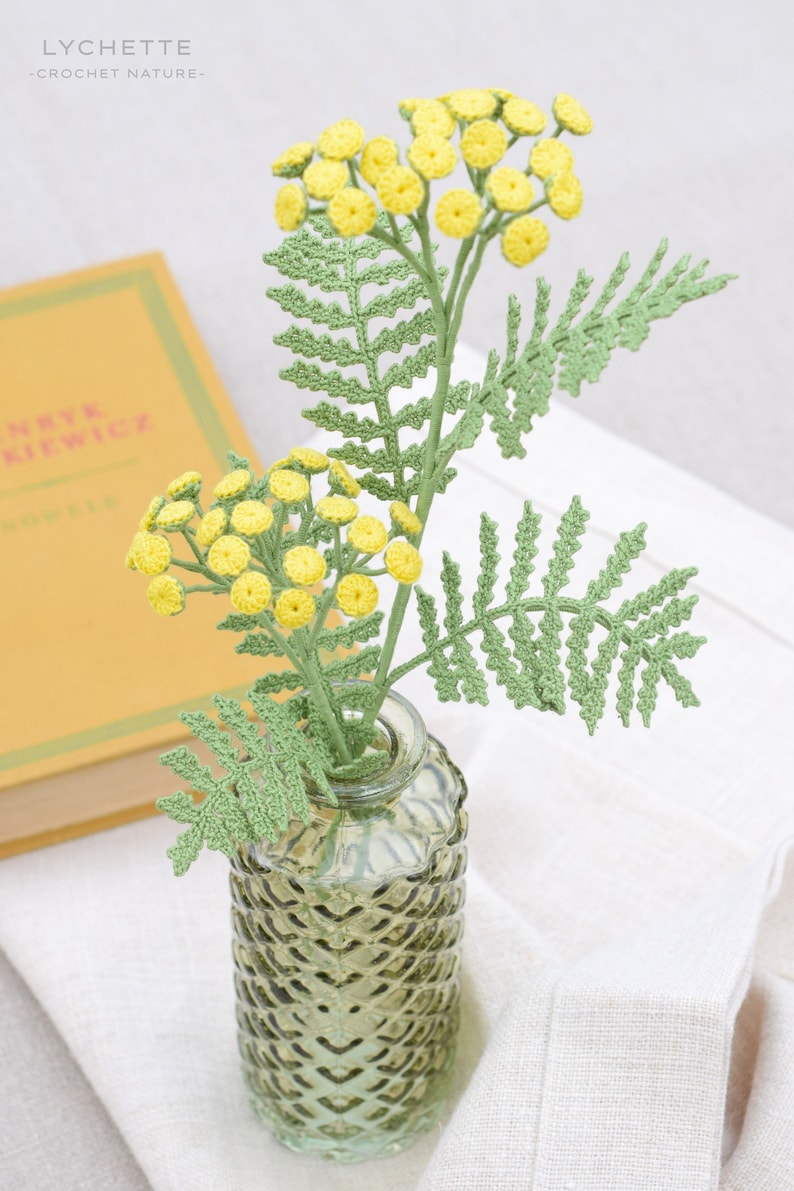 May include: A crocheted bouquet of yellow flowers with green leaves in a clear, textured glass vase. The vase is sitting on a white linen surface.