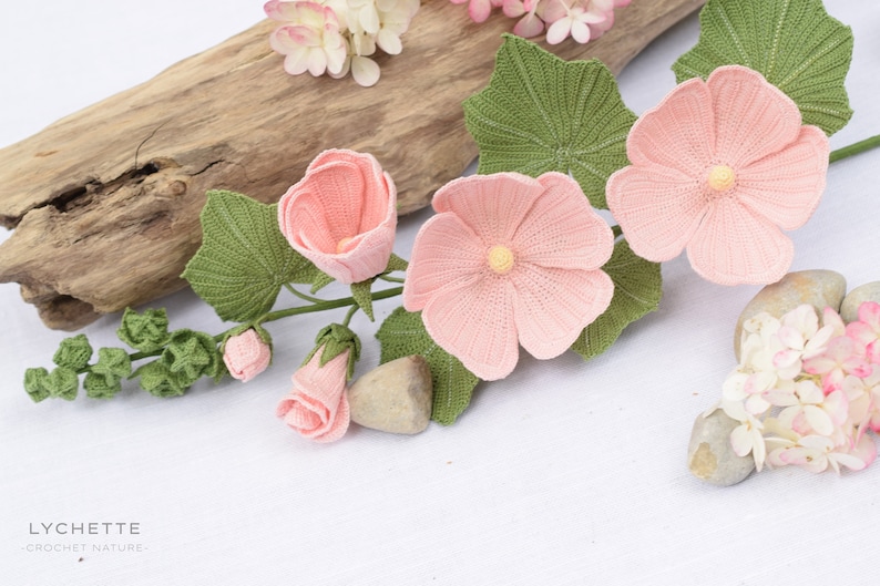 May include: A crocheted flower arrangement featuring pink flowers with green leaves and buds. The flowers are arranged on a light brown wooden surface with a white background. The text "LYCHETTE - CROCHET NATURE -" is visible at the bottom of the image.