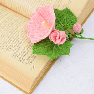 May include: A crocheted bouquet of pink hibiscus flowers with green leaves. The flowers are arranged on an open book.