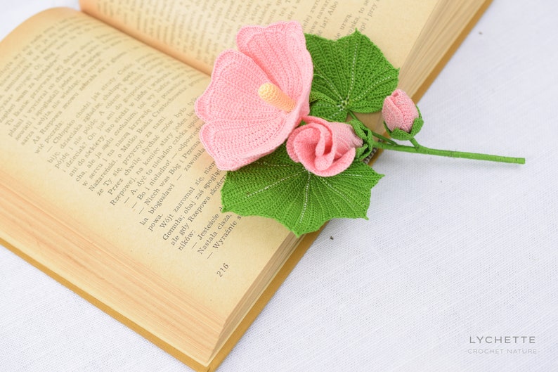 May include: A crocheted bouquet of pink hibiscus flowers with green leaves. The flowers are arranged on an open book.