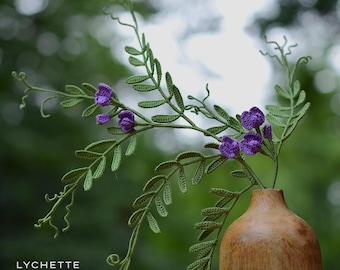 May include: A wooden vase holds a crocheted floral arrangement with purple flowers and green leaves. The vase is a warm brown color, and the crochet work is detailed. The background is a blurred green, suggesting an outdoor setting. The text "LYCHETTE -CROCHET NATURE-" is visible.