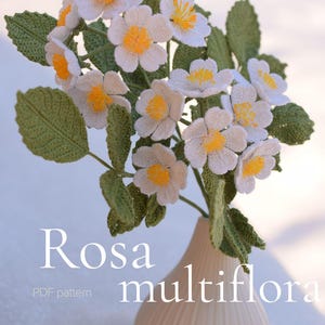 May include: A bouquet of crocheted white flowers with yellow centers and green leaves in a beige vase. The text "Rosa multiflora" and "PDF pattern" are visible. The artist's name, "LYCHETTE", is also displayed.