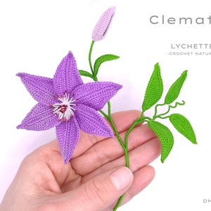 Puede incluir: Una flor de clem&aacute;tide de color p&uacute;rpura hecha a crochet con hojas verdes y un capullo. La flor se sostiene en una mano. El texto "Clematis" y "LYCHETTE -CROCHET NATURE- DMC No 20" es visible en la imagen.
