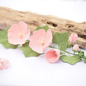 May include: A crocheted flower arrangement featuring pink hibiscus flowers and green leaves. The flowers are arranged on a branch with a few buds. The arrangement is displayed on a white surface with a piece of driftwood behind it.