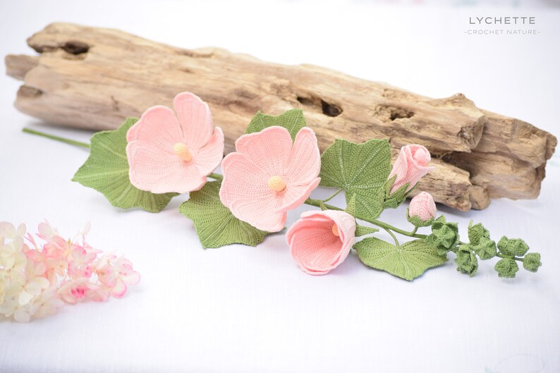 May include: A crocheted flower arrangement featuring pink hibiscus flowers and green leaves. The flowers are arranged on a branch with a few buds. The arrangement is displayed on a white surface with a piece of driftwood behind it.