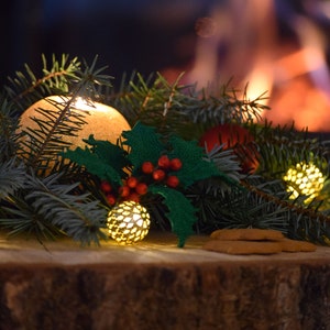 May include: A close-up of a crocheted holly sprig with red berries, surrounded by evergreen branches and lit with small, golden lights. The text "Holly Lychette - Crochet Nature -" is visible above the sprig.