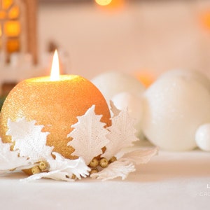 May include: A gold glitter candle with a white crocheted holly design. The candle is surrounded by white ornaments and a pine cone.