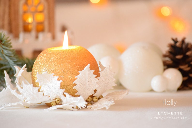 May include: A gold glitter candle with a white crocheted holly design. The candle is surrounded by white ornaments and a pine cone.
