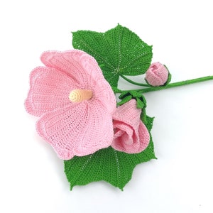 May include: A crocheted pink hibiscus flower with green leaves and buds. The flower is made with a delicate stitch and has a realistic appearance.
