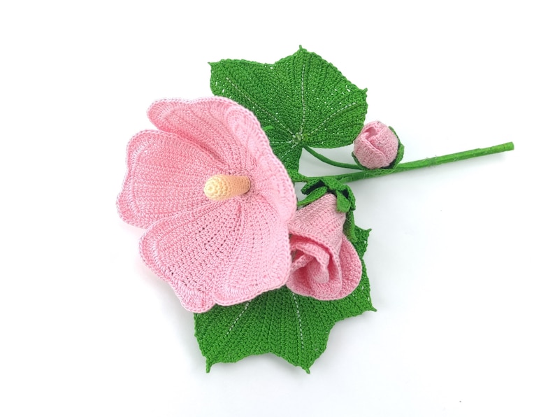 May include: A crocheted pink hibiscus flower with green leaves and buds. The flower is made with a delicate stitch and has a realistic appearance.