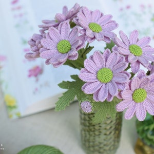 Könnte beinhalten: Ein Strauß gehäkelter lavendelfarbener Gänseblümchen mit grünen Zentren und Blättern in einer strukturierten Glasvase. Die handgefertigten Blumen werden in der Nähe eines Buches und einer grünen Lampe präsentiert. Das Bild ist gut beleuchtet und zeigt die feinen Details der Handarbeit.