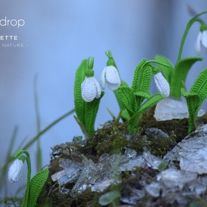 Op de afbeelding: Gehaakte sneeuwklokjes in wit met groene stelen en bladeren. De bloemen zijn gerangschikt op een bed van mos en gebroken ijs.