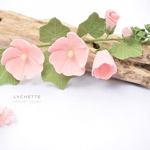 May include: A crocheted flower stem with pink flowers and green leaves. The stem is resting on a piece of driftwood. The text "LYCHETTE -CROCHET NATURE- " is visible below the stem.