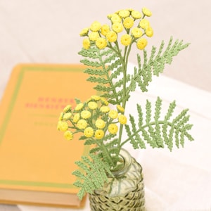 May include: A crocheted bouquet of yellow flowers with green leaves in a green glass vase. The vase is sitting on a book with the text "Henri Matisse" visible.