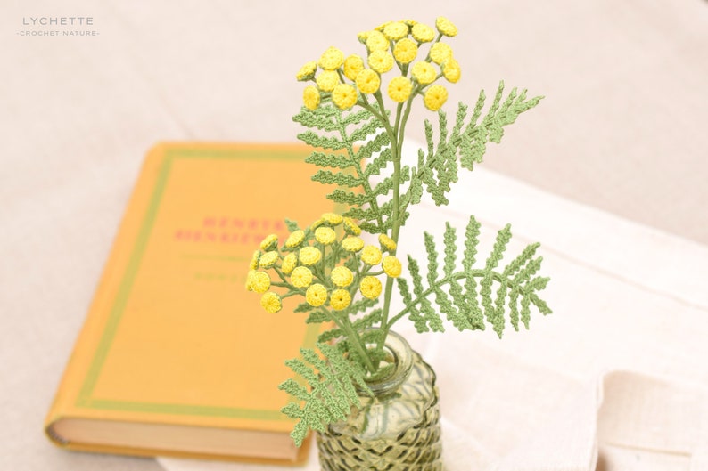 May include: A crocheted bouquet of yellow flowers with green leaves in a green glass vase. The vase is sitting on a book with the text "Henri Matisse" visible.