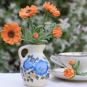 以下が含まれることがあります： 青い花柄の白いピッチャーに、オレンジ色の編み物の花束が活けられています。花は白い表面に置かれた花瓶に活けられています。白いソーサーとティーカップの上に、オレンジ色の編み物の花も置かれています。