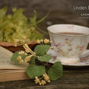 Puede incluir: Una rama de flor de tilo tejida a crochet con delicadas flores amarillas y hojas verdes, descansando sobre un libro y una taza de té y un platillo con diseño floral. La taza de té está llena de té. El texto "Linden Blossom LYCHETTE - CROCHET NATURE -" es visible en la esquina superior derecha de la imagen.