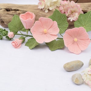 May include: A crocheted flower arrangement featuring pink flowers and green leaves. The flowers are displayed on a white surface with a wooden log and stones in the background.