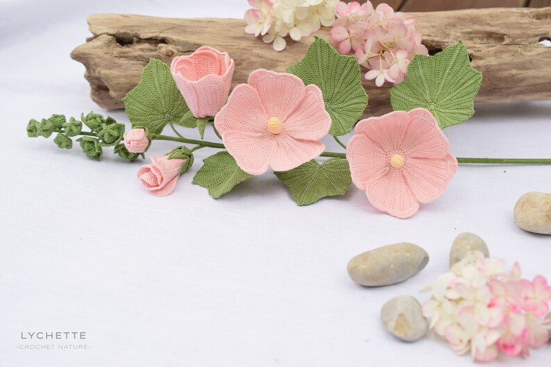 May include: A crocheted flower arrangement featuring pink flowers and green leaves. The flowers are displayed on a white surface with a wooden log and stones in the background.