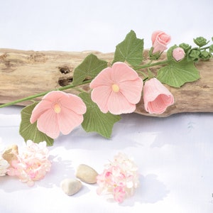 May include: A crocheted pink flower stem with green leaves and buds. The stem is lying on a piece of driftwood. The flowers are delicate and detailed. The background is white.