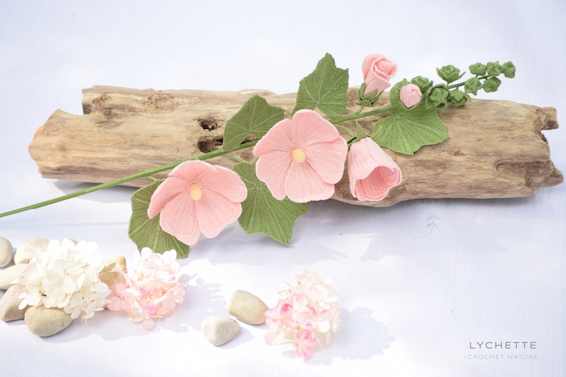 May include: A crocheted pink flower stem with green leaves and buds. The stem is lying on a piece of driftwood. The flowers are delicate and detailed. The background is white.
