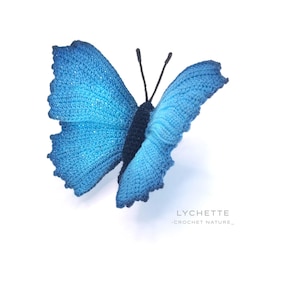 Może przedstawiać: Ręcznie robiony, niebieski motyl z czarnymi czułkami. Motyl znajduje się na białym tle. Pod motylem widnieje napis "LYCHETTE - CROCHET NATURE - ".