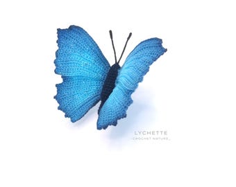 May include: A crocheted blue butterfly with black antennae. The butterfly is on a white background. The text "LYCHETTE - CROCHET NATURE -" is below the butterfly.