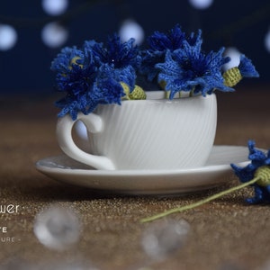 Puede incluir: Una taza de té blanca y un platillo con un ramo de aciano azul tejido a crochet. Las flores están dispuestas en la taza y una flor está en el platillo. El fondo es azul oscuro con luces blancas.