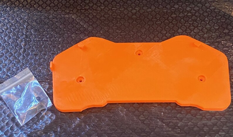 May include: Orange plastic 3D printed part with five screw holes. A small bag of screws is next to the part.
