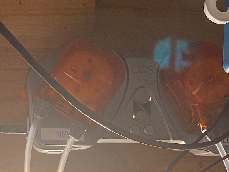 May include: A close-up of a gray and white device with two clear orange pumps. The device has the word "DOS" printed on it. The pumps are connected to clear tubing.