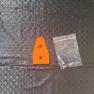 Può includere: Un pezzo di plastica arancione, di forma trapezoidale, con due fori circolari, accanto a un piccolo sacchetto di plastica trasparente contenente diverse piccole viti. Gli oggetti sono su una superficie strutturata grigio scuro.